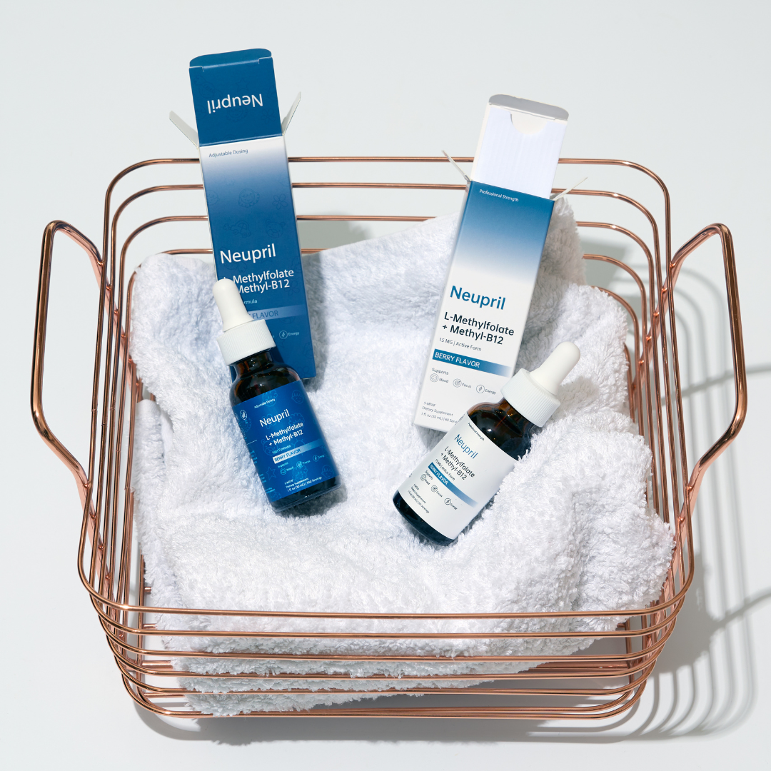 A rose gold wire basket containing a soft white towel, with two Neupril L-Methylfolate + Methyl-B12 dropper bottles and their corresponding blue and white product boxes resting on top. One bottle is the blue Kids Formula and the other is the white Professional Strength version